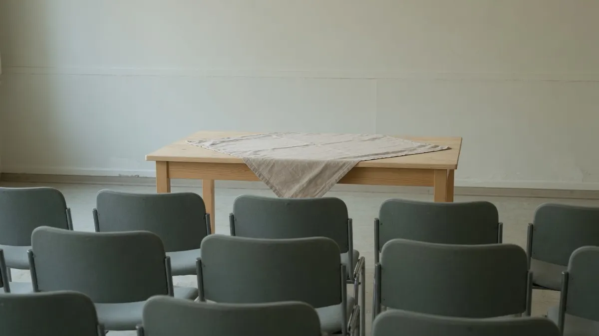 Rzędy szarych krzeseł zwrócone ku jasnemu drewnianemu stołowi nakrytemu lnianym bieżnikiem